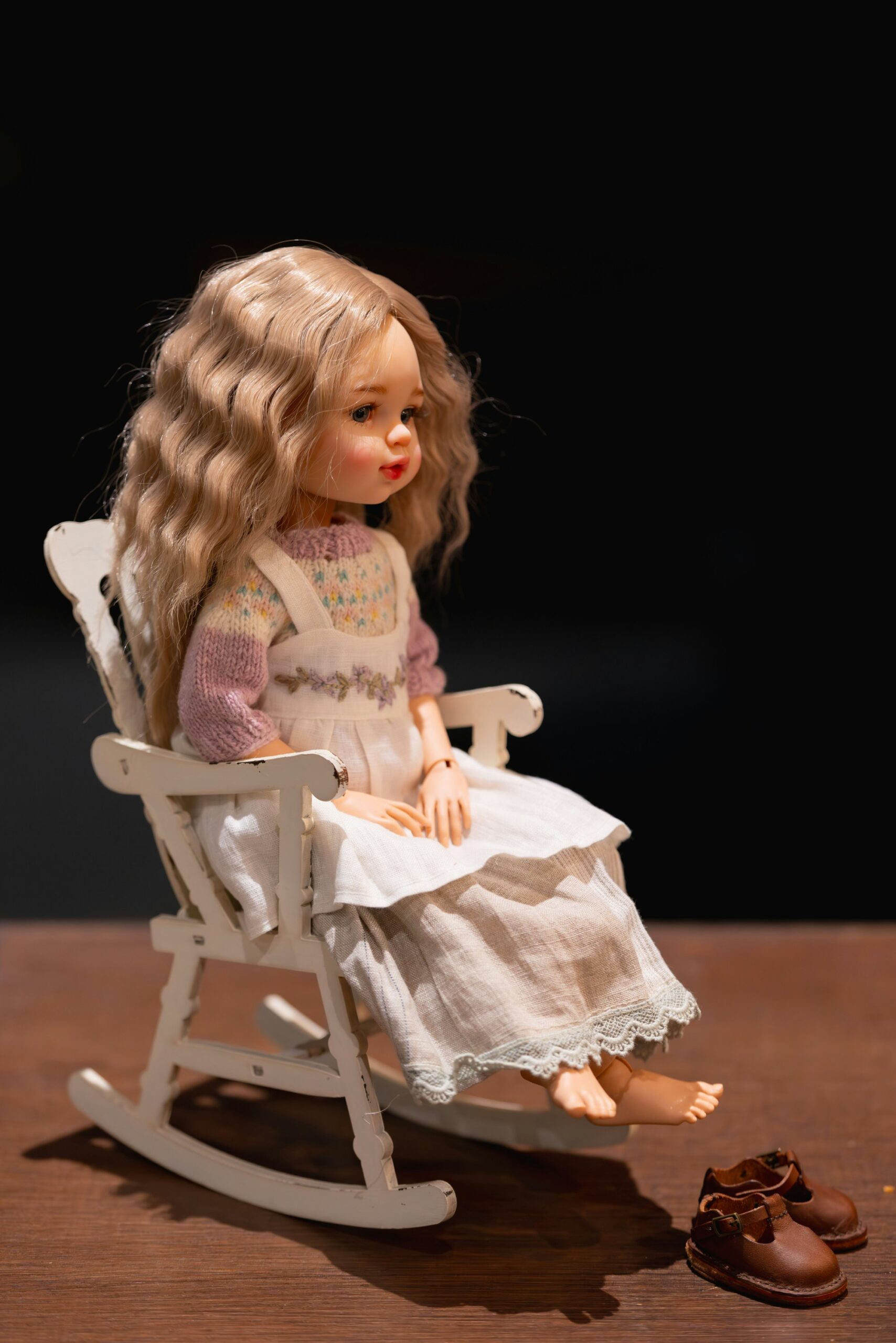 A Blythe-style doll with long wavy blonde hair, wearing a white dress with a knitted pastel sweater, sitting barefoot on a white wooden rocking chair, with a pair of brown doll shoes placed on the floor nearby.